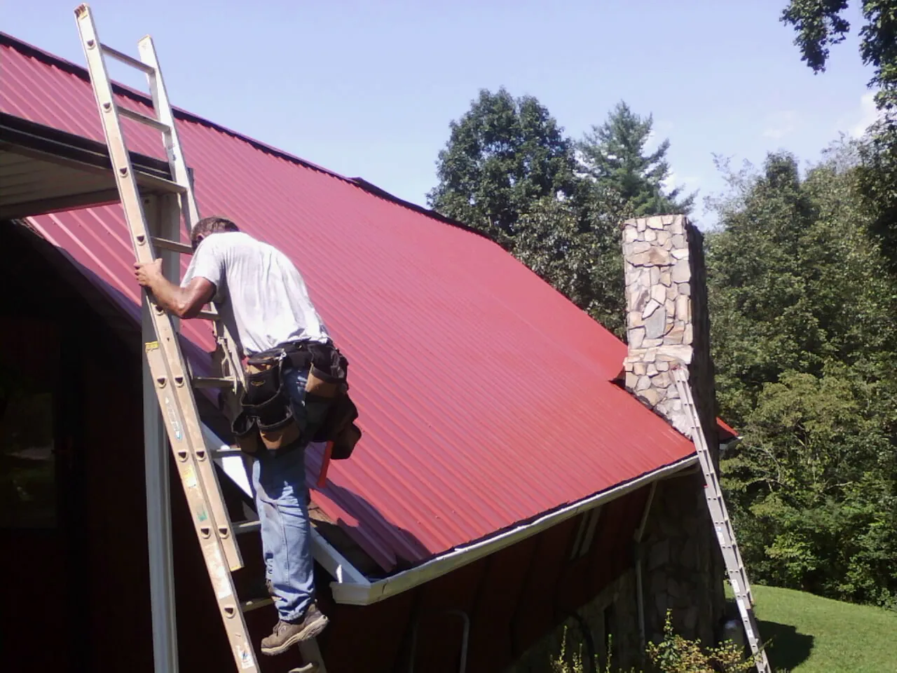 Roofer on a ladder beside a red metal roof and stone chimney — Soffit & Fascia Repair by Roofing Brothers in Colleyville