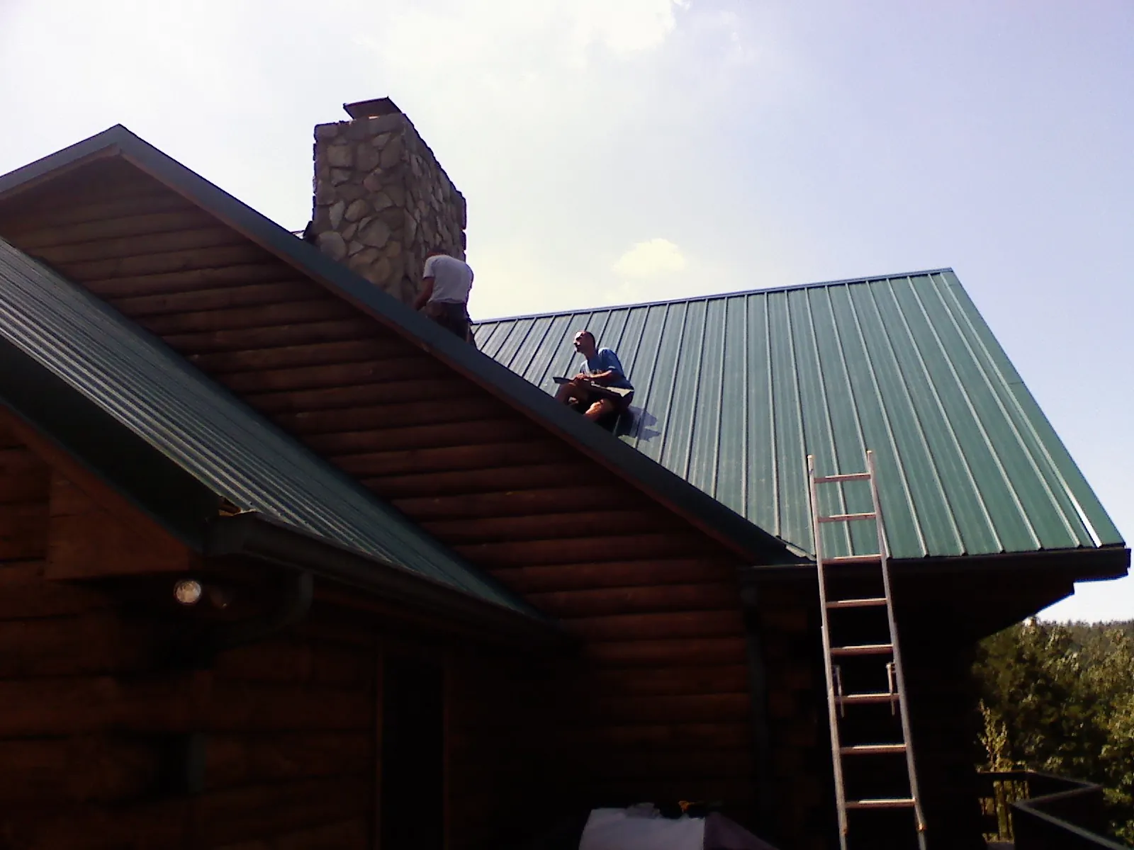Roofers installing green metal panels beside a stone chimney on a log home in Colleyville
